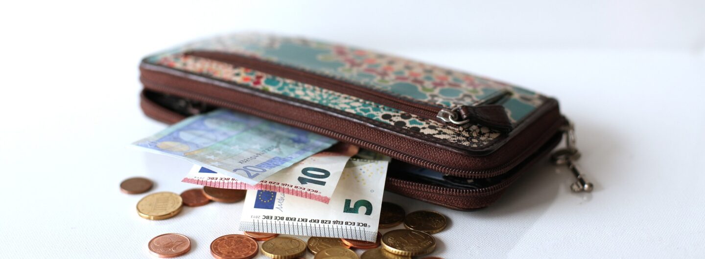 Zu sehen ist ein Portemonnaie mit einigen Euro-Scheinen und Münzen darin und daneben.