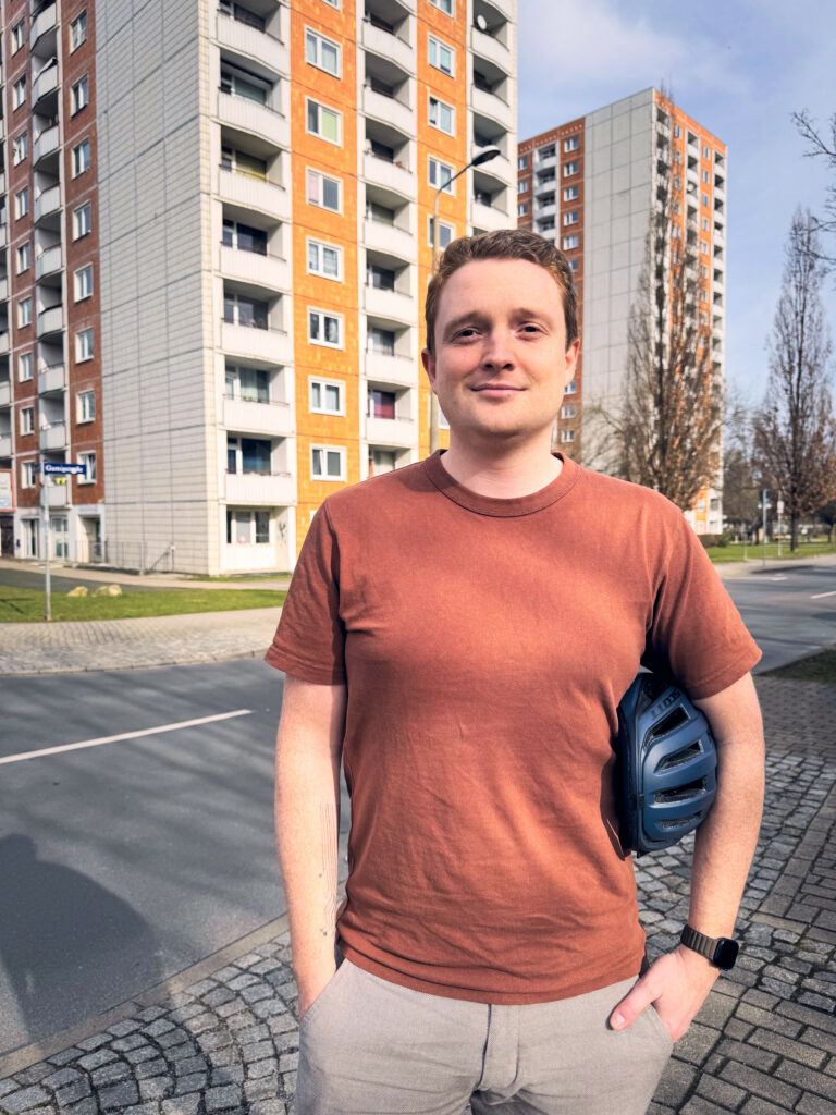Kandidat Kilian Költzsch, der mit einem Fahrradhelm unter dem Arm vor einer Straße steht, im Hintergund ein Plattenbau.