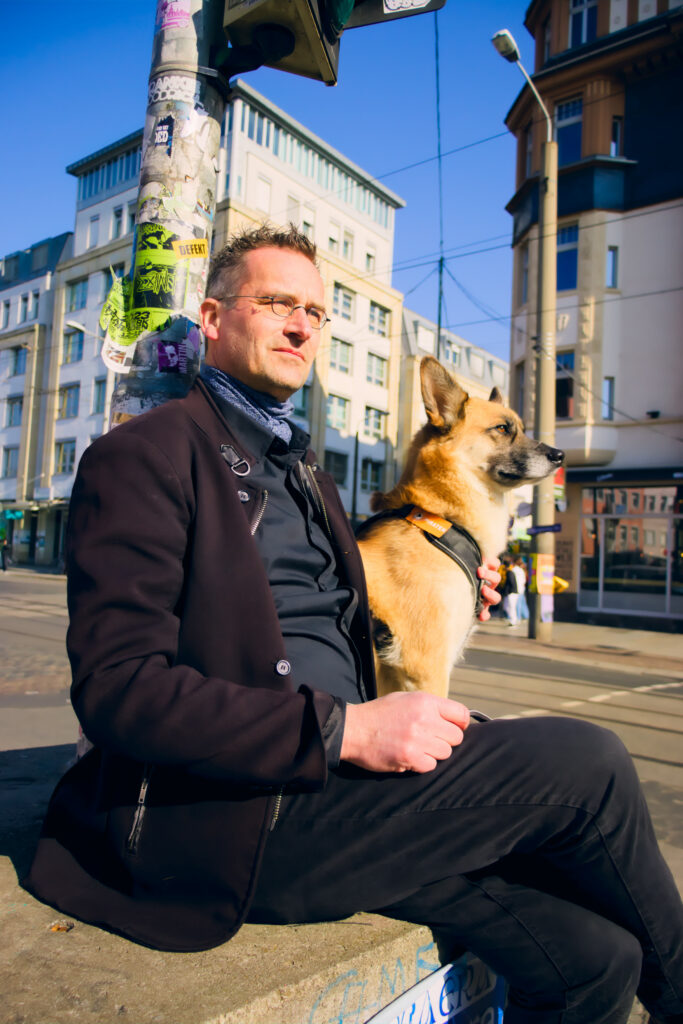 Kandidat Martin Schulte-Wissermann, der mit seiner Hündin auf dem Betonblock einer temporären Ampel vor einer Straße mit Häusern sitzt.