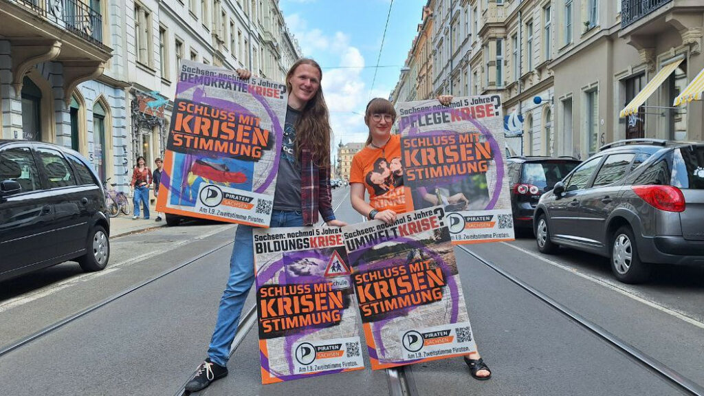 2 Personen auf einer Straße zeigen lächelnd 4 Wahlplakate in die Kamera.