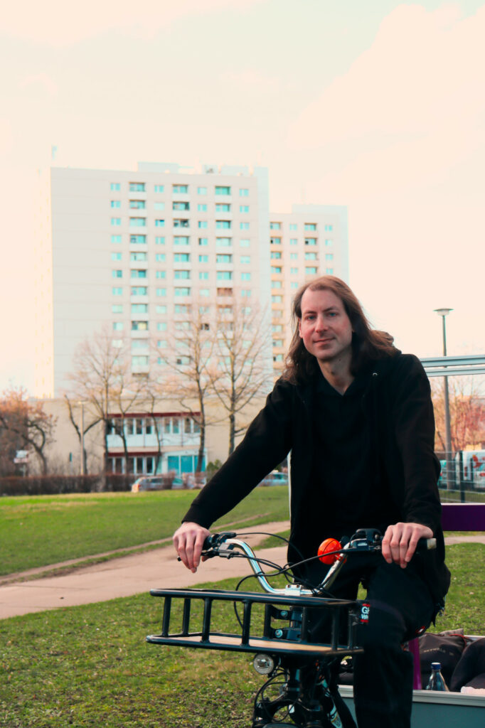 Kandidat Daniel Quitt, der auf einem Fahrrad in einem Park sitzt, im Hintergrund ein Plattenbau.