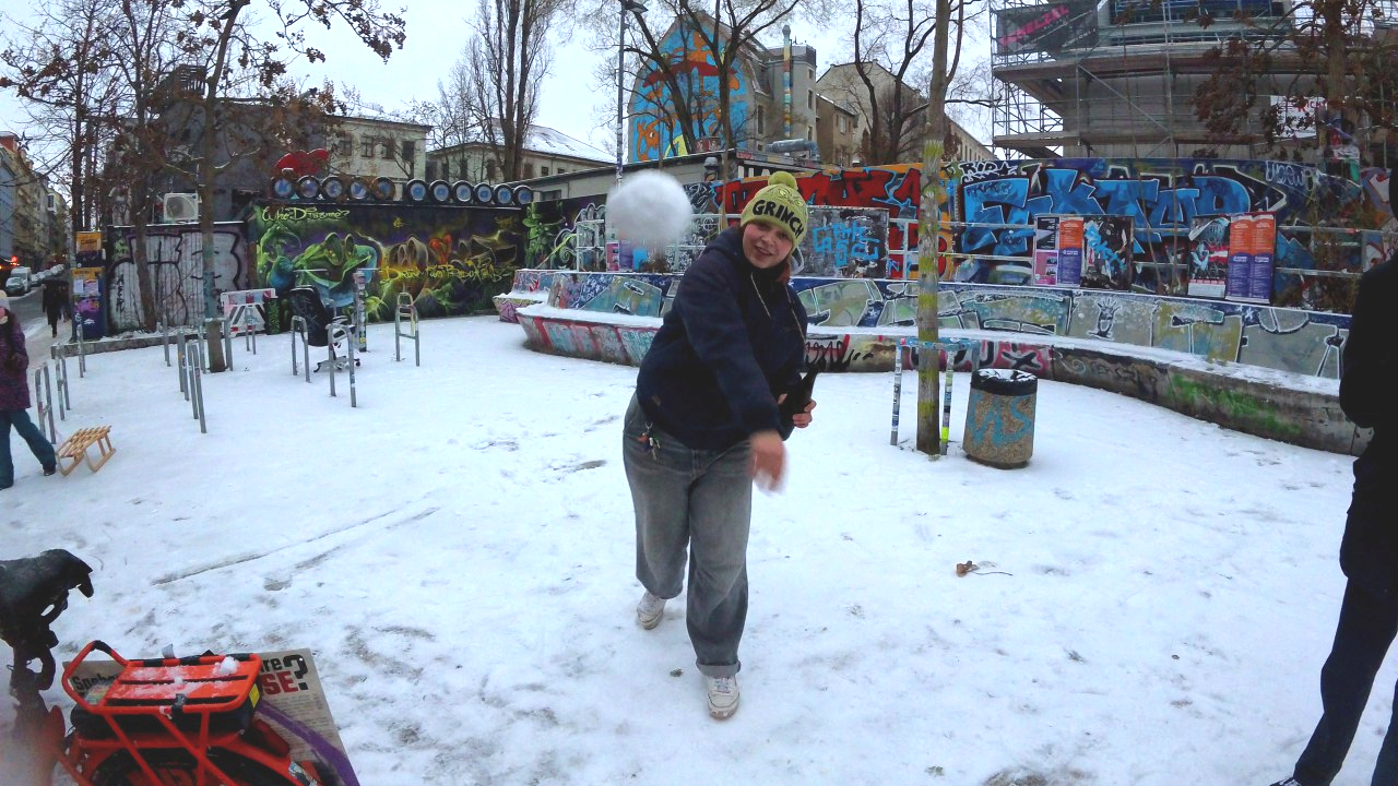 Eine Person wirft einen Schneeball in die Kamera. Im Hintergrund ist ein buntes Stadtviertel mit Bäumen und bemalten Wänden zu erkennen.