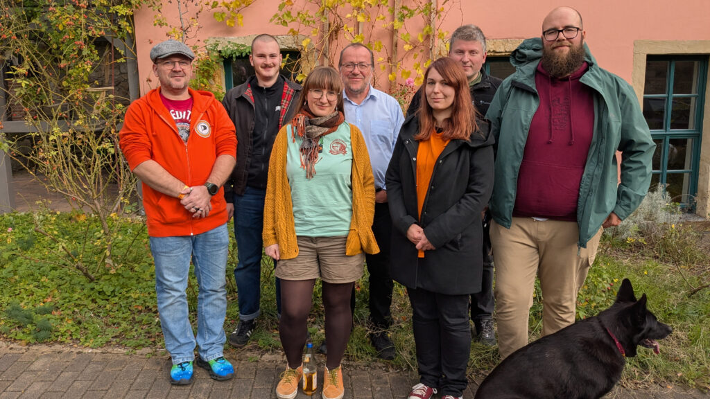 Sieben Personen vor einer bewachsenen Hauswand mit einem Hund.
