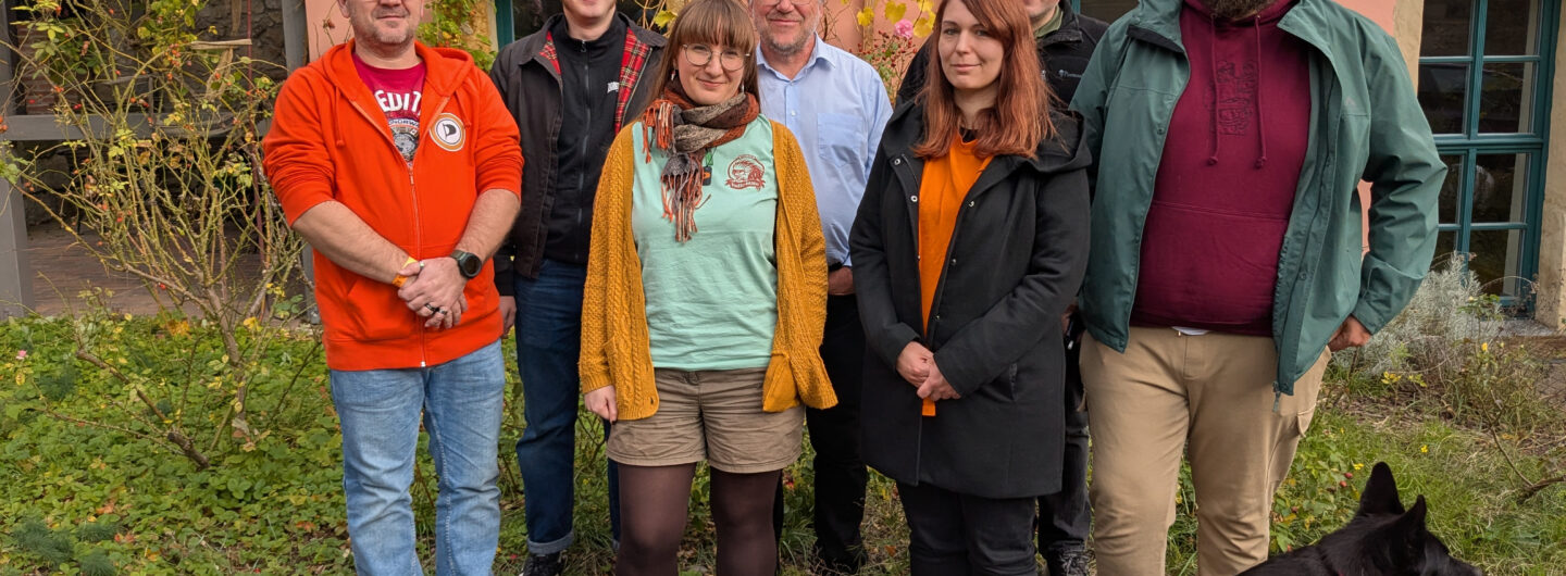 Sieben Personen vor einer bewachsenen Hauswand mit einem Hund.