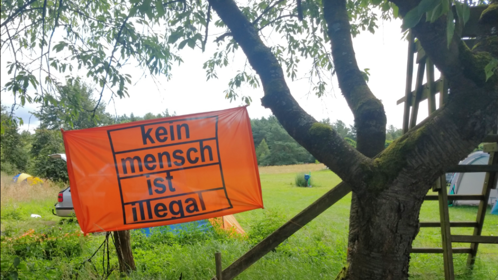Eine orange Flagge an einen Baum gespannt auf der: "Kein Mensch ist illegal" steht.