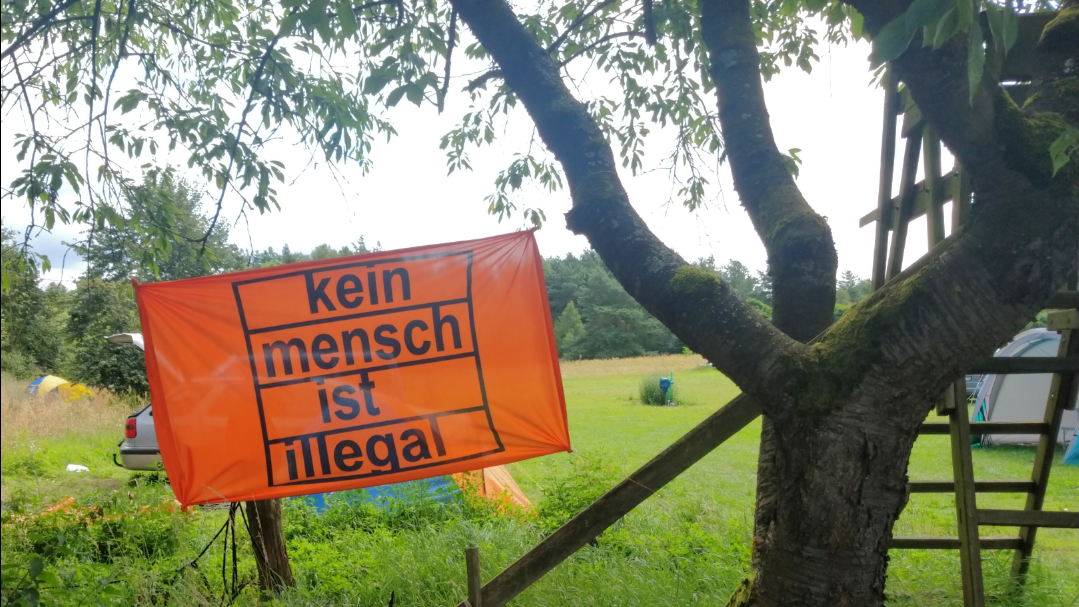 Eine orange Flagge an einen Baum gespannt auf der: "Kein Mensch ist illegal" steht.